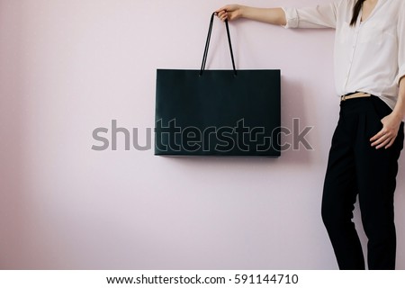 Young hipster girl wearing white blouse and black trousers and holding blank black shopping bag, mock-up of black paper package, pink modern background
