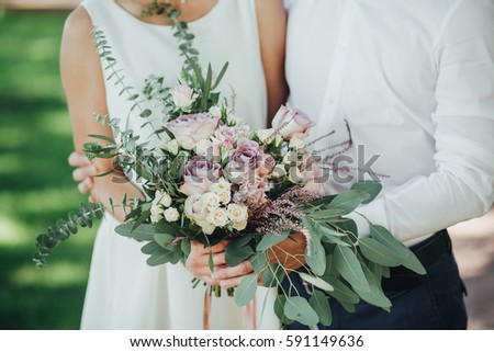 bride and groom in a light white dress standing on the old wooden stairs in the forest and are holding wedding bouquet of flowers and greenery