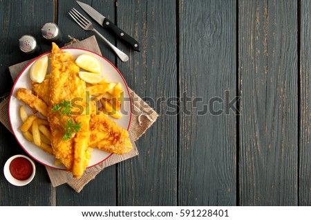 Two pieces of battered fish on a plate with chips on a wooden table with space 