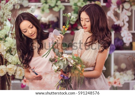 Beautiful asian florist girls making bouquet of flowers for sale against floral bokeh background in flower shop indoors. Two attractive asian females florists working in retail store. 2 playful