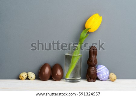 Chocolate bunny and eggs, a tulip, quail and Easter eggs on the grey background, close-up. Light wooden surface. Happy Easter.