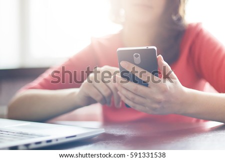 Film effect.  A shot of young female dressed in red casual sweater using high-speed Internet connection on cell phone sitting at the table near her laptop. People, technology and lifestyle concept.