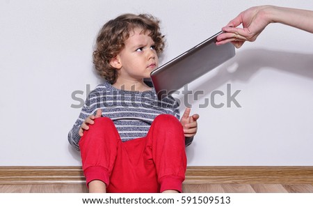 Beautiful kid playing games on a tablet. Boy sitting on the floor and looking on the display of a tablet watching cartoons. Very angry kid because his mother took the tablet from him