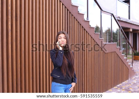 Cute girl with long hair cute talking on phone with friend or loved one, make an appointment. Women dressed in black sweater, blue jeans, black boots on platform and top dressed in jacket standing on
