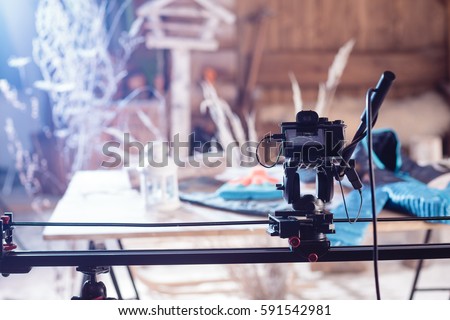 Detail of the camera on the slider and tripod in front of blurred colorful background with plants, bird feeder and wooden wall. Moment from shooting with blue color filter.
