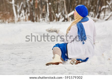 Plump smiling woman in skates sits in snowdrift and catches snowflakes on mitten at outdoor skating rink in winter park.