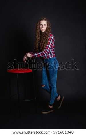 studio portrait of beautiful thin curly young woman teen girl, isolated on black background posing  in a red plaid shirt and tight jeans. standing near a red bar stool