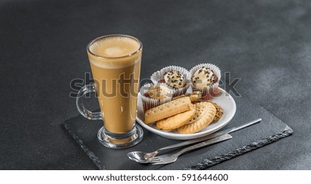 left side view on an aromatic coffee latte with three buttered muffins and petit fours on a white saucer with a spoon and forks, the whole on a black stone plate, sweet set