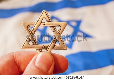 Fingers holding a Star of David (Magen David), Jewish traditional symbol, hexagram with a flag of Israel on the background. Jewish diaspora. Israel Independence Day. Conversion to judaism concept Royalty-Free Stock Photo #591968147