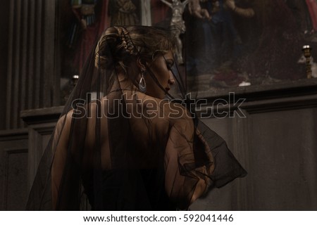 young blond woman with beautiful hairstyle is covered by a black veil in black dress kneeling at the altar in the background of paintings and arches and prays to Jesus