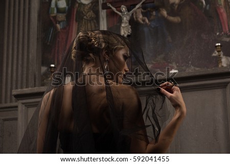 young blond woman with beautiful hairstyle is covered by a black veil in black dress kneeling at the altar in the background of paintings and arches and prays to Jesus looking away