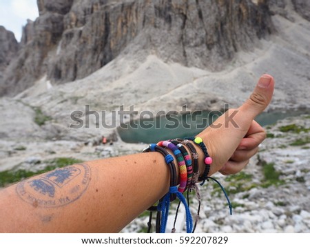 Hand with bracelets, stamp (of Al Pisciadu Hutte) and thumb up in front of lake and mountains in Dolomites, Italy
