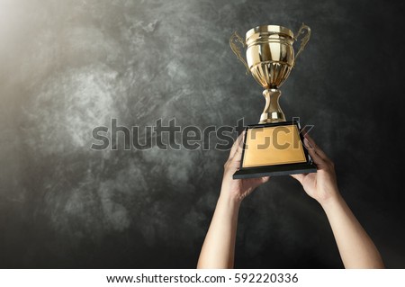 a man holding up a gold trophy cup with grunge wall background copy space ready for your trophy design. Royalty-Free Stock Photo #592220336