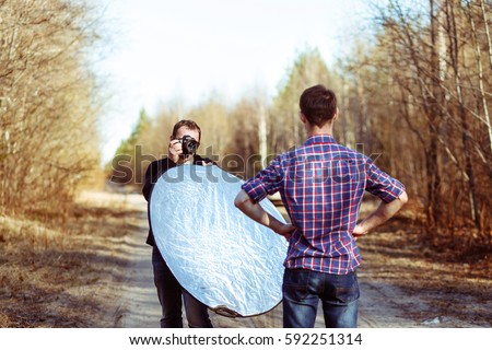 Photographer Photographing Male Model in Forest. Backstage of Fashion Photoshoot by Professional Photographer with DSLR Camera and Reflector