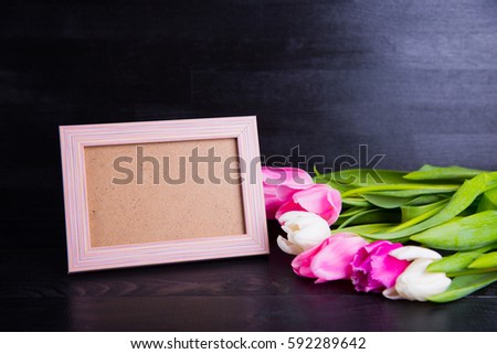 Beautiful bouquet of tender pink and white tulips and empty photo frame on black wooden background. Spring romantic composition.