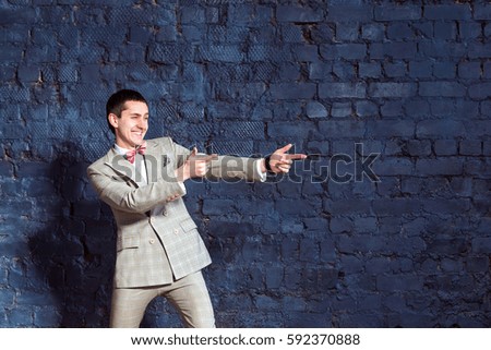 Elegant smiling show man dressed in suit is pointing at blank space at blue wall background.