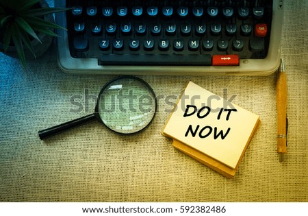 DO IT NOW wording on notebook with typewriter ,pen and 
magnifying glass on wooden table. Motivation and positive wishes concept