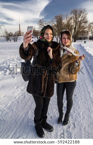 Two young and attractive women are taking a picture of themselves, standing in the park, dresses in fur and painted shawls.
