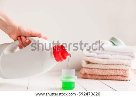 Close up of female hands pouring liquid laundry detergent into cap on white rustic table with towels on background in bathroom. Royalty-Free Stock Photo #592470620
