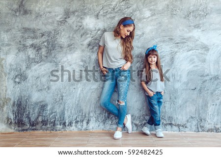 Young beautiful mom with her daughter wearing blank gray t-shirt and jeans posing against rough concrete wall, minimalist street fashion style, family same look, clothing for parent and child. Royalty-Free Stock Photo #592482425
