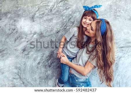 Young beautiful mom with her daughter wearing blank gray t-shirt and jeans posing against rough concrete wall, minimalist street fashion style, family same look, clothing for parent and child. Royalty-Free Stock Photo #592482437