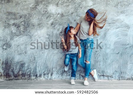 Young beautiful mom with her daughter wearing blank gray t-shirt and jeans posing against rough concrete wall, minimalist street fashion style, family same look, clothing for parent and child. Royalty-Free Stock Photo #592482506