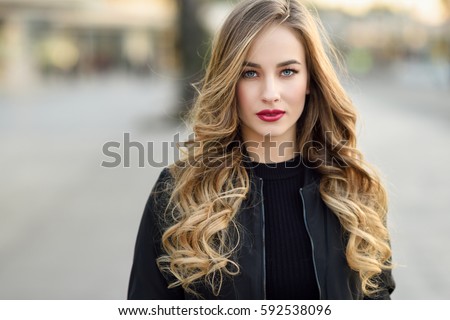 Close-up portrait of young blonde girl with beautiful blue eyes wearing black jacket outdoors. Pretty russian female with long wavy hair hairstyle. Woman in urban background.  Royalty-Free Stock Photo #592538096