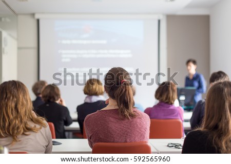 Female speaker giving presentation in lecture hall at university workshop. Rear view of unrecognized participants listening to lecture and making notes. Scientific conference event. Royalty-Free Stock Photo #592565960