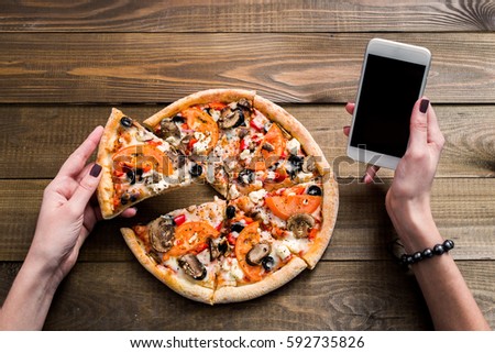 hands of a woman ordering pizza with a device over a wooden workspace table. All screen graphics are made up. Royalty-Free Stock Photo #592735826