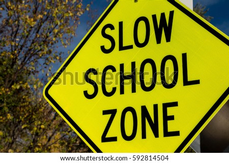 Yellow caution sign that says Slow School Zone against autumn tree and sky