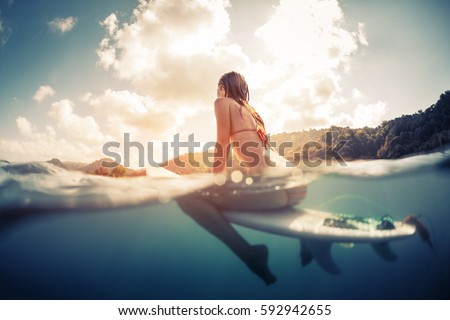 Split shot of the young woman sitting on the surfboard in the ocean with green sunrise hills on the background
