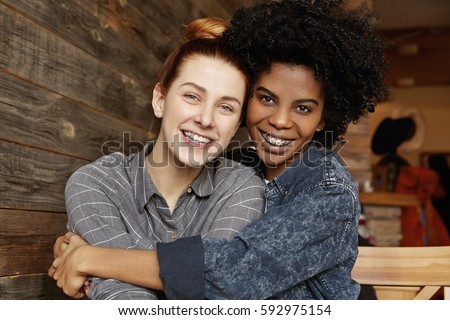 Sweet tender indoor shot of happy interracial homosexual couple hugging and cuddling at cafe: African-American fashionable girl holding tight her charming Caucasian girlfriend. People and relationship Royalty-Free Stock Photo #592975154