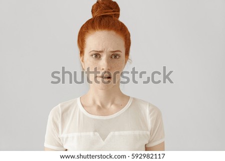 Confused or puzzled beautiful young Caucasian woman with ginger hair frowning, biting her lower lip after having done something wrong, looking at camera with guilty and apologetic facial expression Royalty-Free Stock Photo #592982117