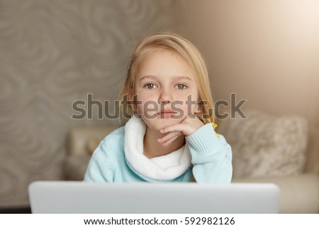 Indoor portrait of adorable cute European little girl with fair hair using laptop computer in her room, watching cartoons and video blogs online, enjoying leisure time after classes at school