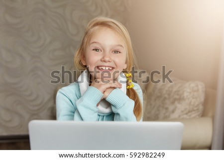 Happy cute little girl with messy ponytail smiling happily, resting face on hands, watching cartoons online on generic laptop. European female child using wireless internet connection on notebook pc