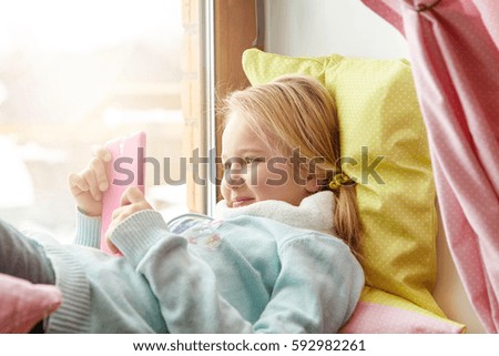 Cheerful little girl in blue sweater lying on windowsill with pillows, happy with using new mobile phone, bought by parents on her birthday, enjoying wireless internet connection, messaging friends