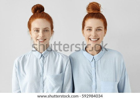 Two young redhead Caucasian females looking alike wearing same formal light-blue shirts, looking at camera, smiling happily, standing close to each other against white studio wall background Royalty-Free Stock Photo #592984034