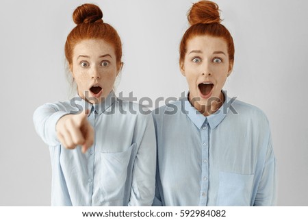 Two beautiful Caucasian females looking alike with hair buns, wearing same formal shirts, opening mouths widely, having surprised shocked looks, one of girls pointing finger at camera. Selective focus Royalty-Free Stock Photo #592984082
