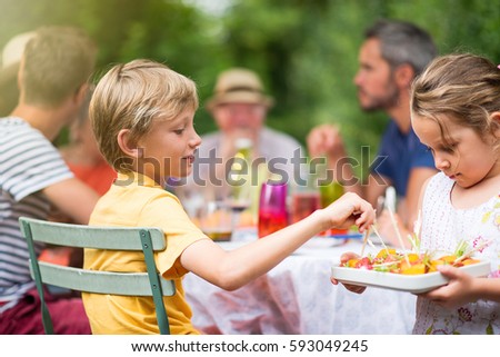 Three generation family having lunch in the garden in summer. A little girl serving a dish with melon and tomatoes. Shot with flare