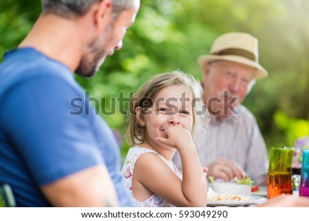 Three generation family gathered to lunch in the garden in summer, focus on a beautiful little girl looking at camera. Shot with flare