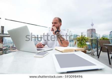 Handsome male prosperous CEO checking correspondence on email box writing feedbacks to partners suggesting collaboration while resting in rooftop cafe using modern digital gadgets and  free wifi