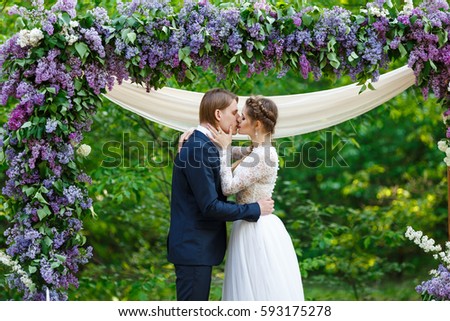 Young beautiful groom in dark blue suit and bride in white crop top dress standing near flower arch with lilac and long ribbons and kissing. Happy couple on spring wedding ceremony Royalty-Free Stock Photo #593175278