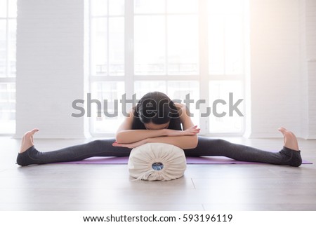 Beautiful young yoga girl lying in asana Shavasana or Corpse Pose using bolster. Fitness woman resting after working out, doing yoga exercise near a window on gym