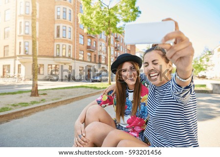Candid image of a laughing young woman taking a selfie with her trendy young friend as they sit together on the curb in a quiet urban street