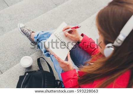 young smiling beautiful hipster happy woman in city street, writing in diary, hands close-up, details, listening to music on headphones, spring trend, urban teenage style,  Royalty-Free Stock Photo #593228063