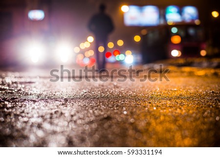 Rainy night in the big city, glare from the headlights of the car parked and people passing near. Close up view from the sidewalk level