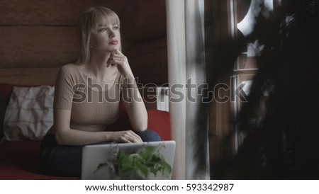 Young pensive woman sitting on the couch while typing and reading emails on the laptop. 