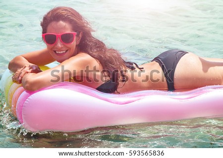 Portrait of young Caucasian woman wearing black bikini and sunglasses lying on pool raft in water, looking at camera and smiling. Female tourist resting at seaside during vacation