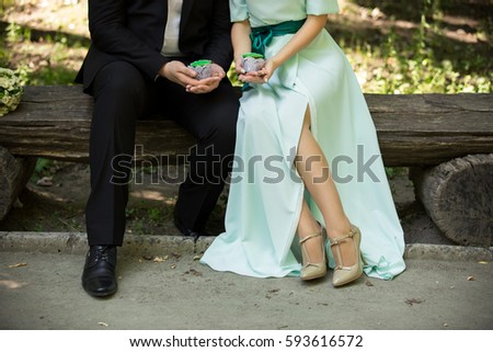 Man and woman holding green cupcakes in park. Green wedding or saint patrick day concept. Stylish couple, bride in mint dress and shoes on high heels, groom in black suit. Wedding accessories