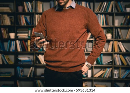 The young man, a businessman in a brown sweater and shirt holding a smartphone. It should be against the background of a cabinet with books in the library or office